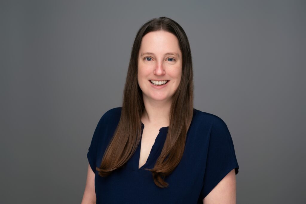 Jen headshot wearing a navy blue blouse against a grey background.