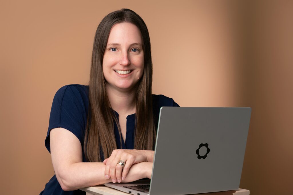 Photo of Jen seated with her laptop. Jen is wearing a navy blue blouse against a brown background.